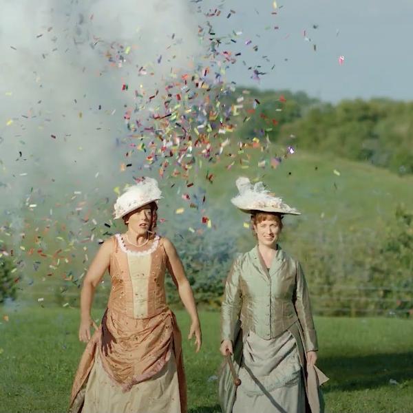 Two women, dressed in 1800s clothes, stand in a field in-front of an explosion of confetti.